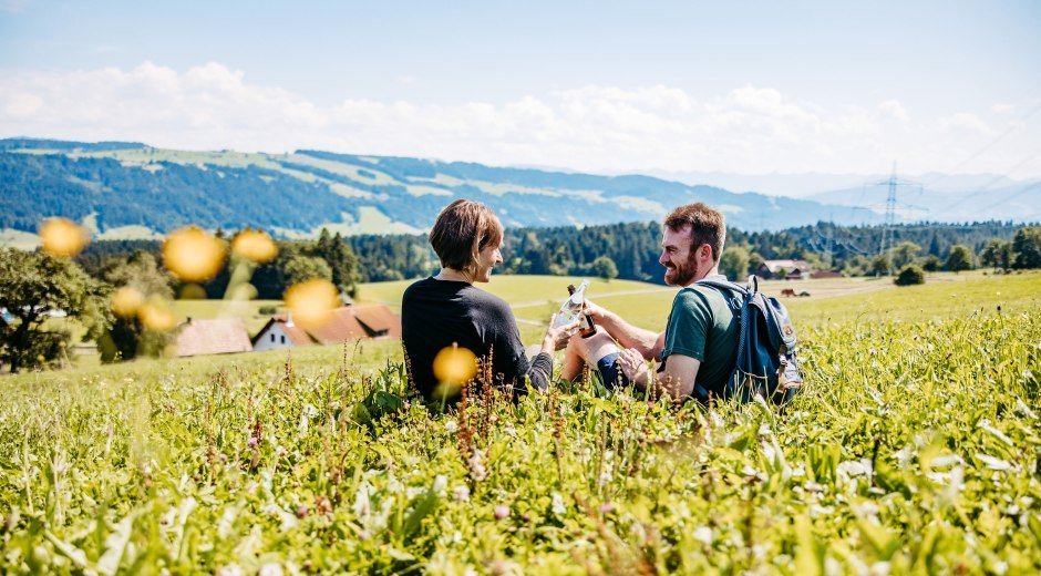 Zwei Personen sitzen auf einer Wiese und trinken Wasser und Bier aus der Region Westallgäu und genießen dabei die Aussicht.  © Frederick Sams